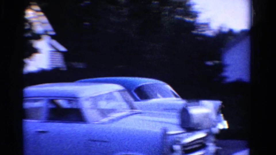 Blue-tinted 1958 Ontario 8mm frame with a white sedan in motion near residential houses and trees.