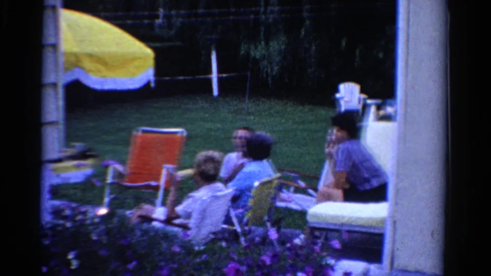 1958 Ontario backyard home-movie frame with adults relaxing on lawn chairs near a yellow umbrella and flowers.