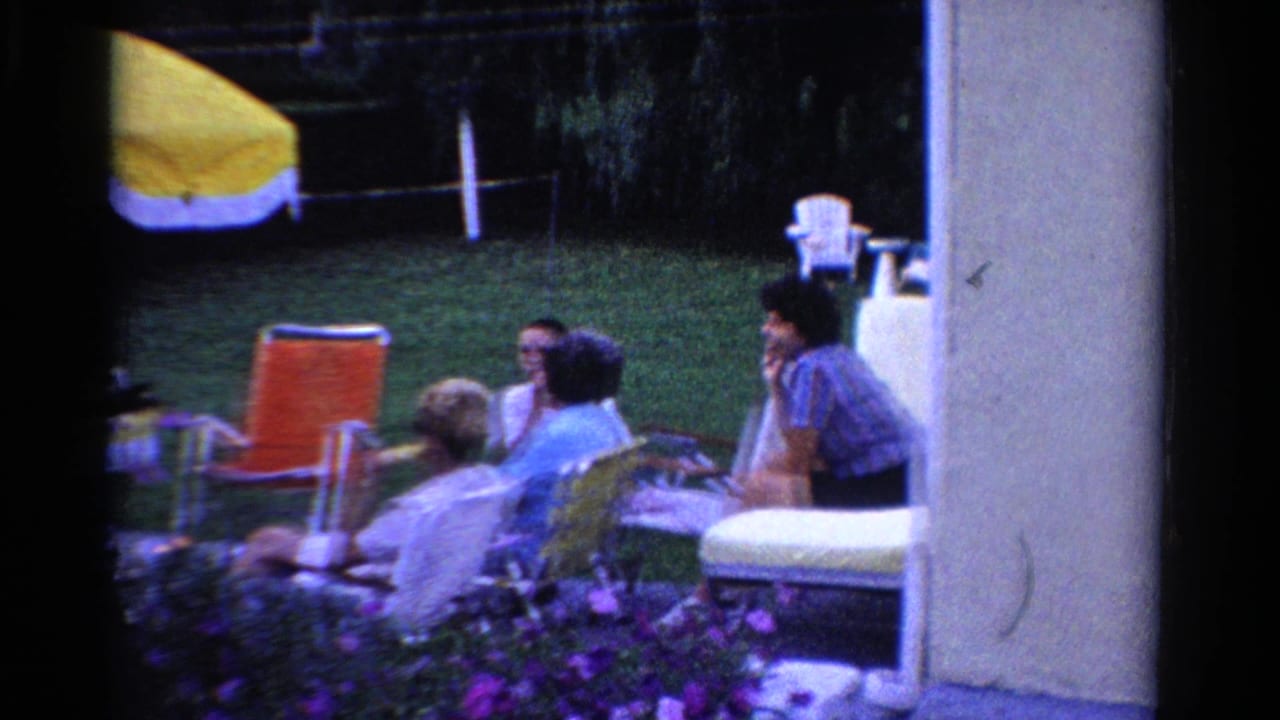 Seated relatives watch from lawn chairs near a yellow umbrella and patio furniture.