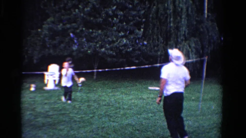 Two adult men play badminton on a green Ontario backyard lawn in 1958, with one player wearing a white cowboy hat and holding a racket.