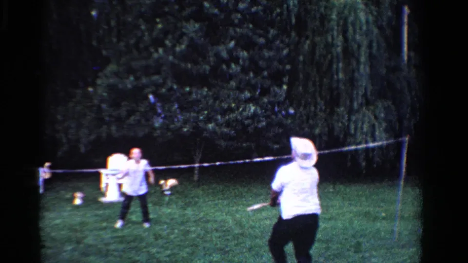 In the 1958 Ontario home movie, two men continue a backyard badminton game, framed by trees, lawn chairs, and a stretched net.