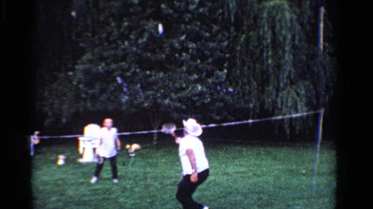 1958 Ontario home-movie frame showing two people playing badminton on a lawn with trees and a white outdoor chair behind them.