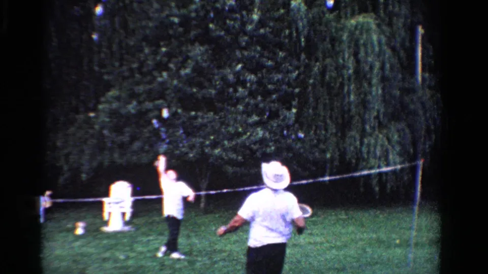1958 Ontario backyard badminton scene with a white-hatted player in the foreground and another player near the net.