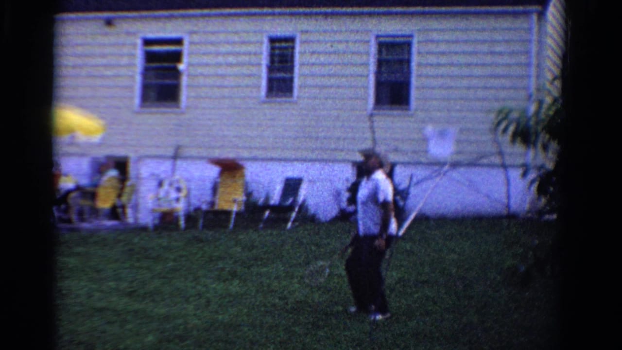 1958 Ontario home-movie frame with an adult badminton player in a backyard near a light-sided house and seated guests.