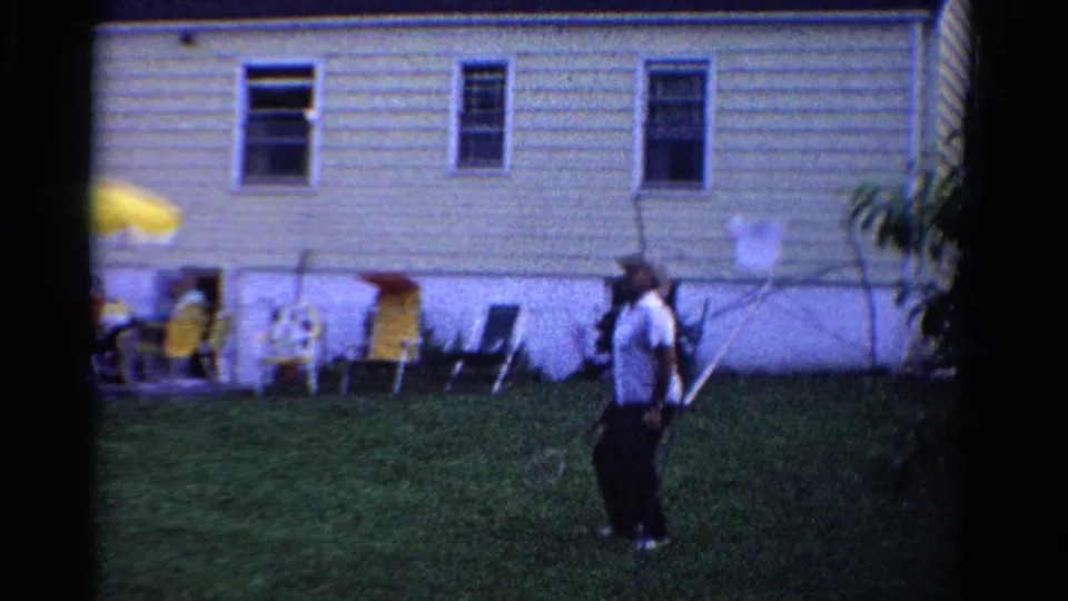 1958 Ontario home-movie frame with an adult badminton player in a backyard near a light-sided house and seated guests.