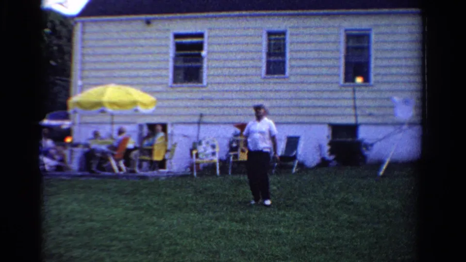 In this 1958 Ontario home-movie frame, a backyard player stands in front of patio chairs, seated guests, and a light-colored house.