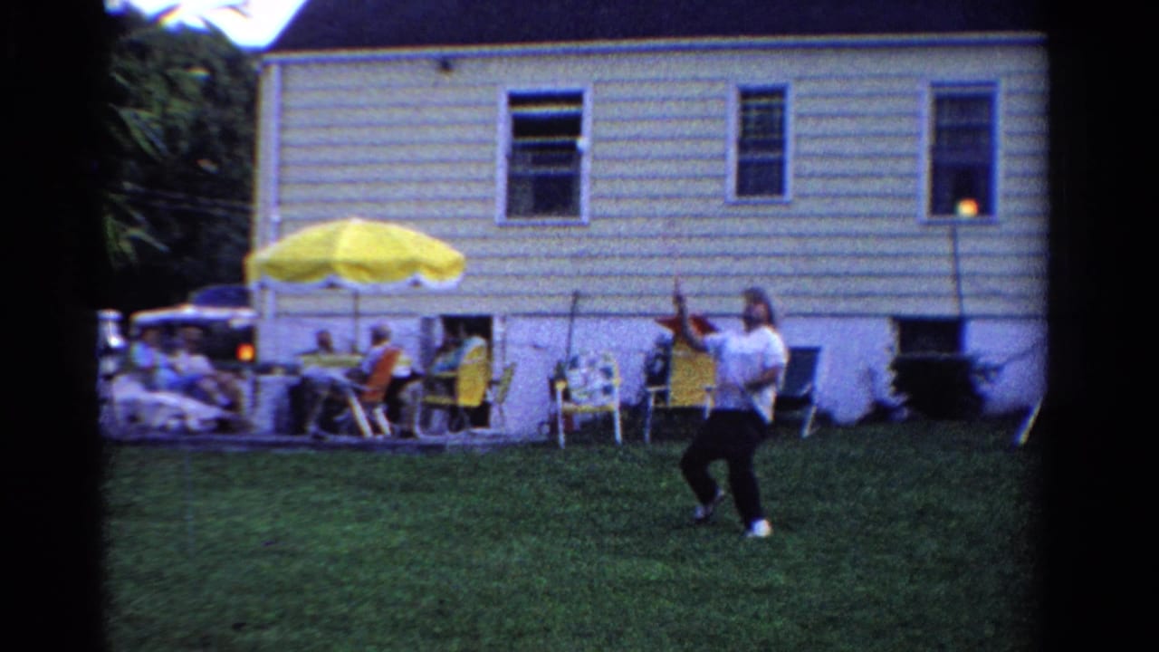 Man swings a badminton racket on a backyard lawn while people sit under a yellow umbrella near a house.