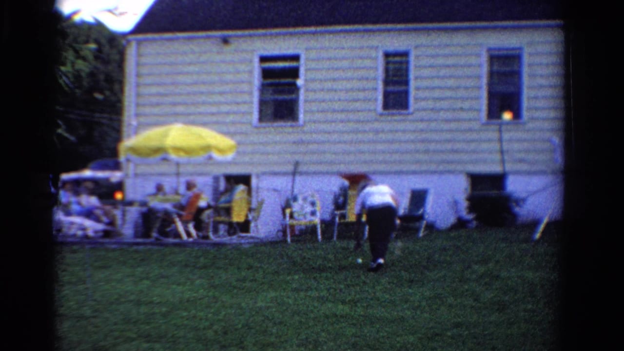 A backyard badminton moment in Ontario, Canada, shows the player bending near the lawn while family members sit by the house under a yellow umbrella.