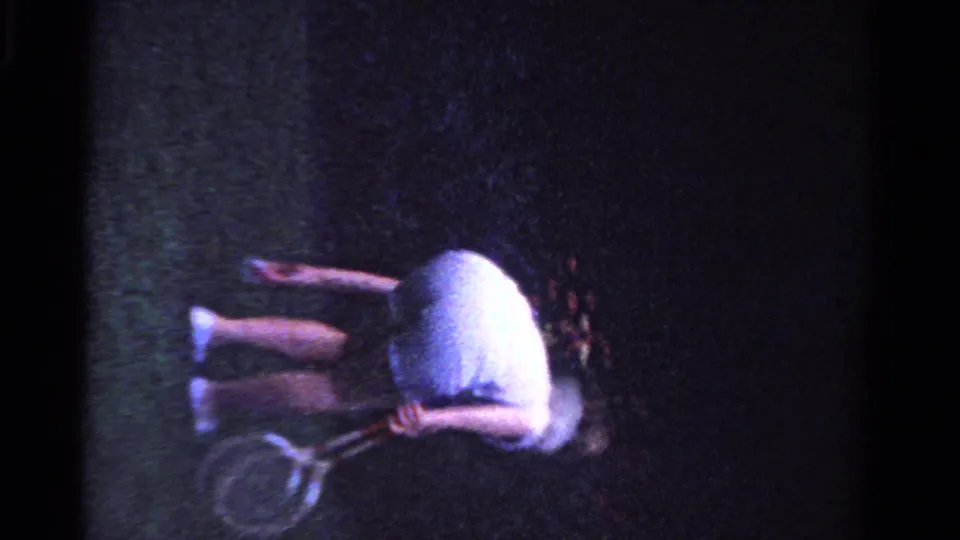 A tilted home-movie frame shows the badminton player low on the lawn beside his racket, with soft focus and dark edge falloff from the 8mm scan.