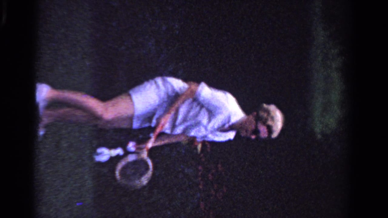 In 1958 Ontario home-movie footage, a woman in white tennis clothing and sunglasses bends forward holding a racket on grass.