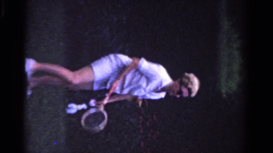 In 1958 Ontario home-movie footage, a woman in white tennis clothing and sunglasses bends forward holding a racket on grass.