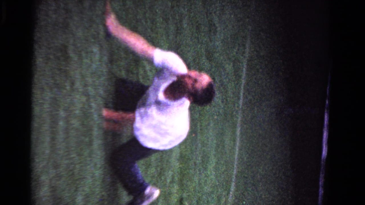 Young man in a white shirt swings a tennis racket on a dark grass court.