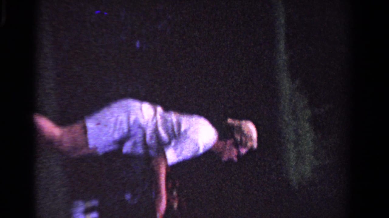 A young man appears mid-movement on a dim grass tennis court in 1958 Ontario home-movie footage.