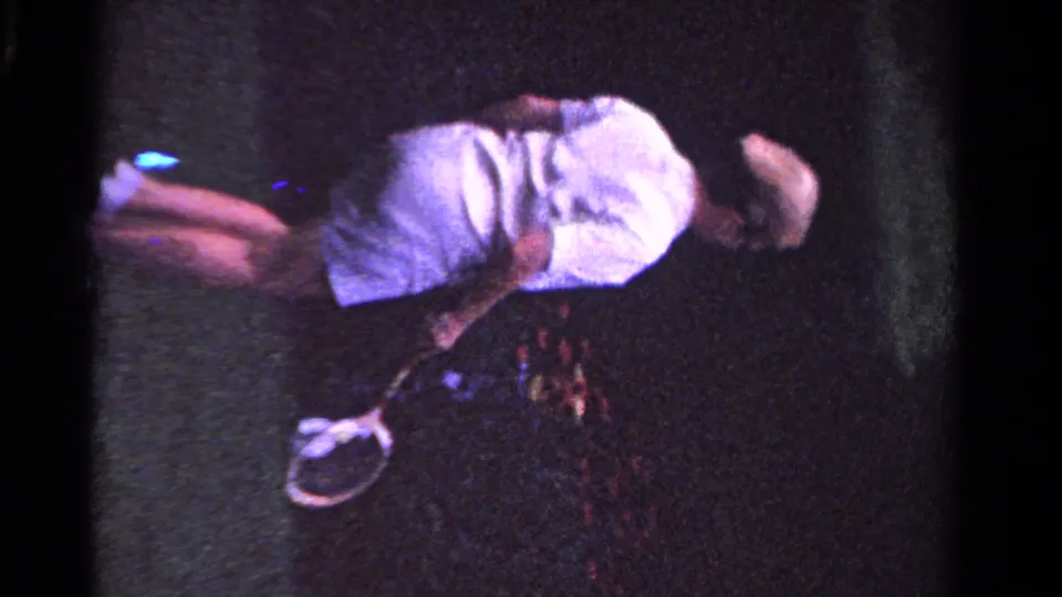 The player bends low with a tennis racket on a dark grass court, captured in grainy 1958 Ontario 8mm footage.