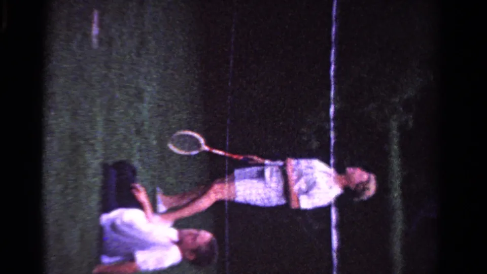 1958 Ontario home-movie frame showing a man with a racket on a grass tennis court and a family member lying nearby.