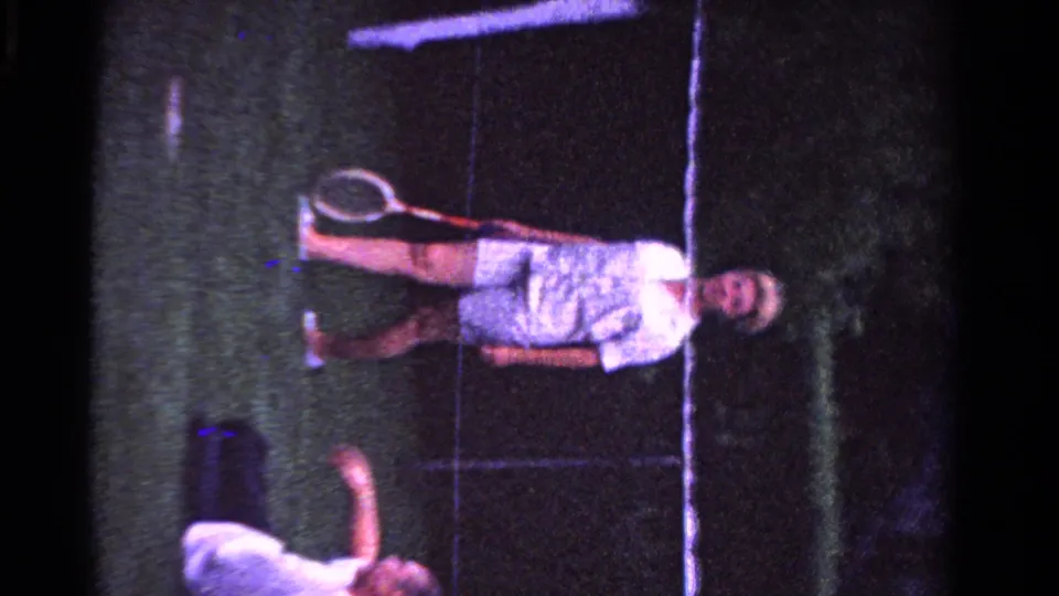 1958 Ontario frame of a man standing on a grass tennis court with a racket while another family member is partly visible below.