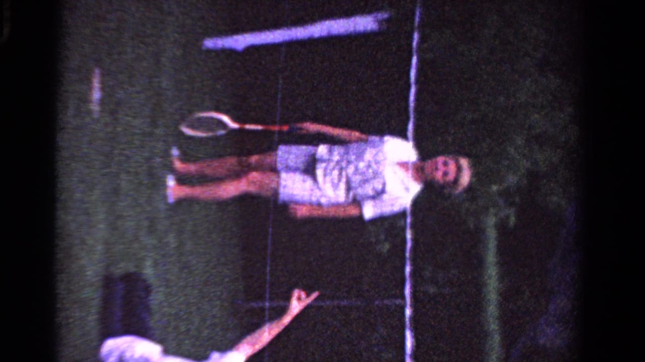 Woman standing on grass with a tennis racket while another person reaches up in the foreground.