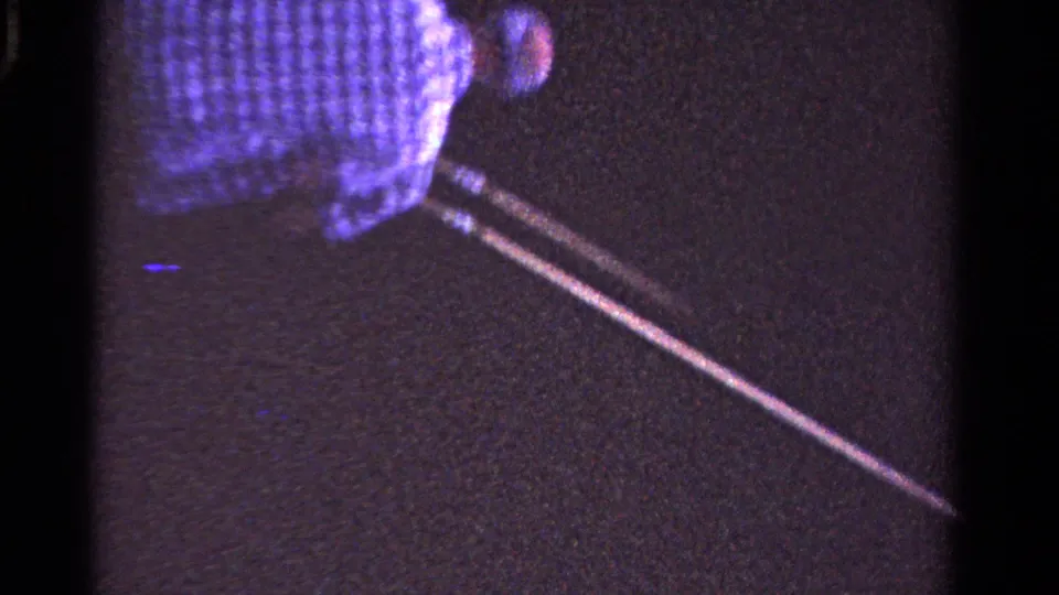 A man in a checkered shirt moves across the grass near a long pole-like object, seen from a tilted handheld 8mm viewpoint.