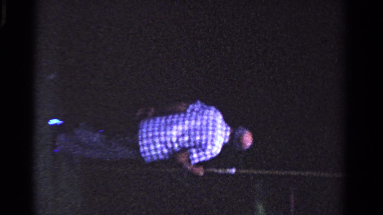 Man in checkered shirt crouches with a long pole in a dark outdoor scene.