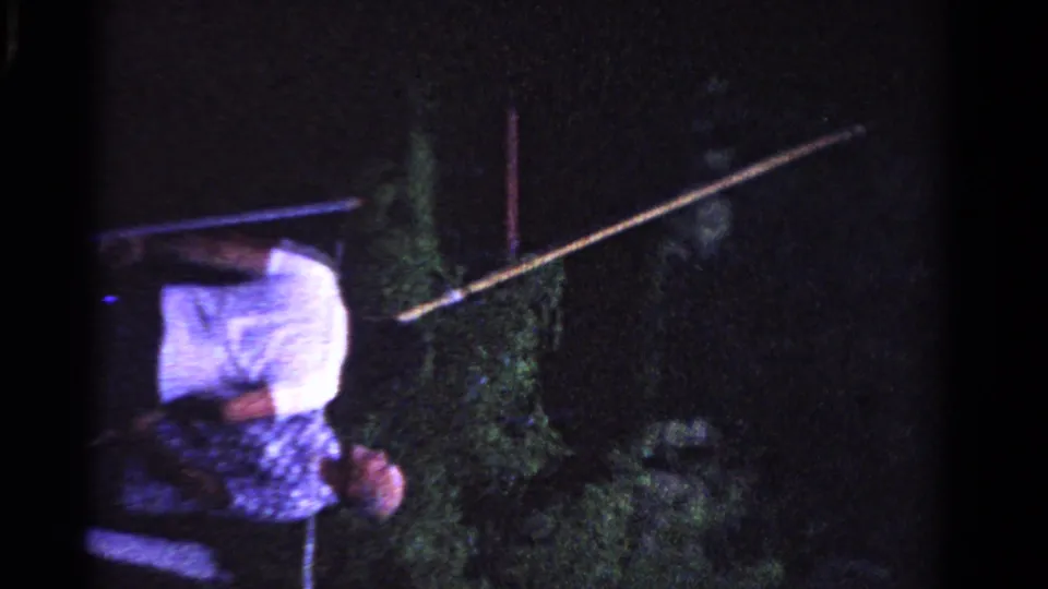 The man angles a long pole upward toward faint foliage in low-light 8mm footage from Ontario, Canada in 1958.