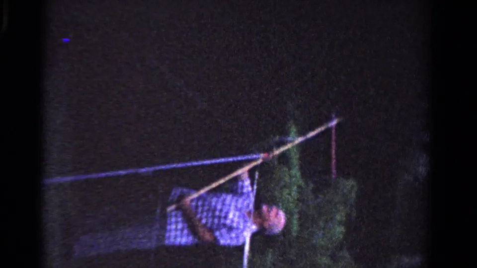 A man in a checkered shirt stands near faint foliage, holding a long pole upward in grainy 1958 Ontario home-movie footage.