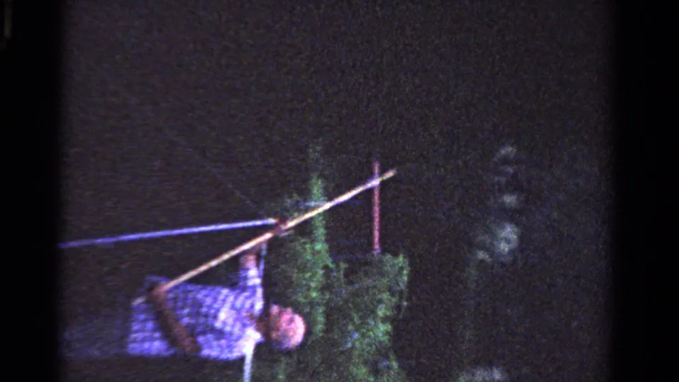 In 1958 Ontario home-movie footage, a man in a blue check shirt leans toward foliage while holding a long pole against a dark sky.