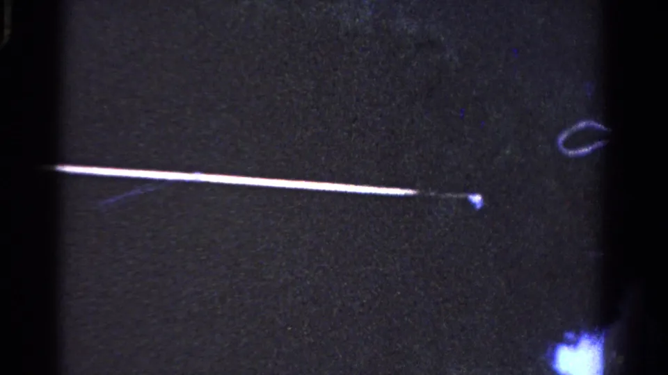 A long pole stretches through the dark 1958 Ontario home-movie frame, ending near a small bright point and looped wire-like form.