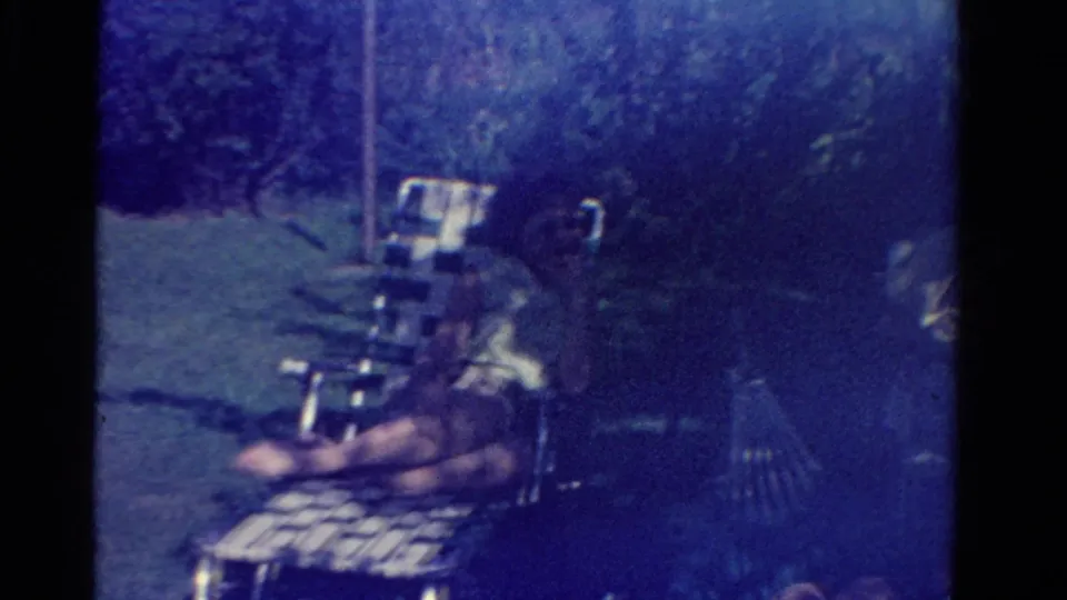 In this Ontario 1958 home-movie frame, a person in a white shirt sits on a striped lawn chair beside grass and trees.
