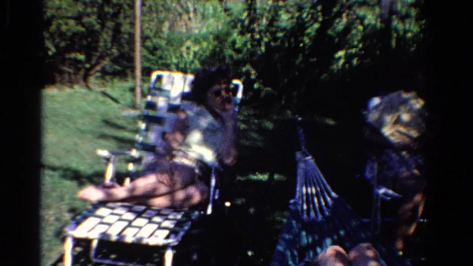 1958 Ontario home-movie frame of adults seated in a backyard, with metal lawn furniture, grass, trees, and a hammock in view.