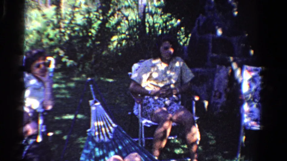 1958 Ontario home-movie frame showing adults seated in a backyard, one near the left edge and one centered beside a hammock.