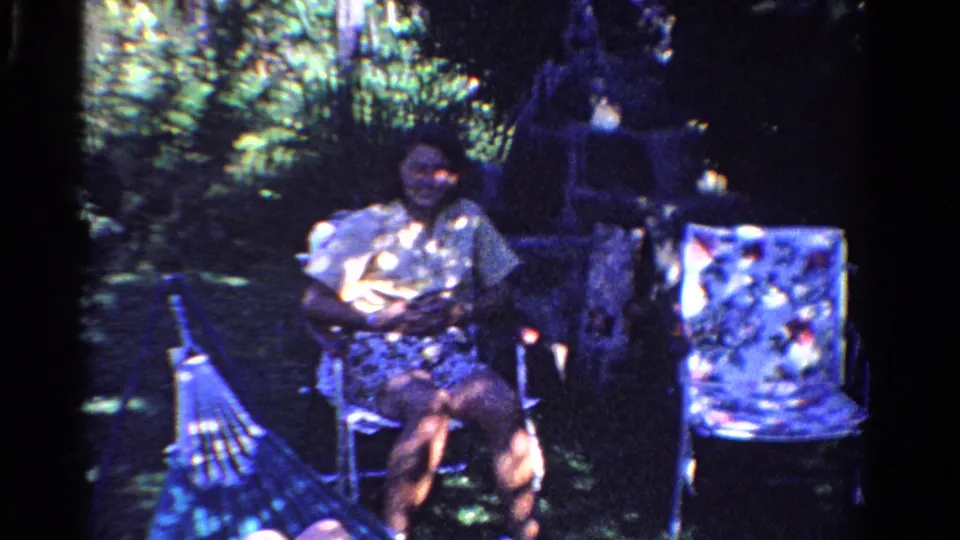 1958 Ontario backyard frame with a seated woman, hammock, lawn chair, grass, and dense trees in soft 8mm color.