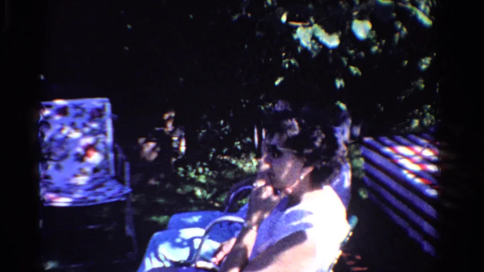 The backyard gathering continues with a woman seated close to the camera, a patterned chair, and leafy garden shade visible.