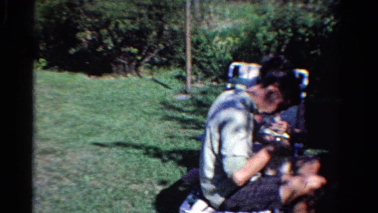 1958 Ontario home-movie frame showing a person in a folding lawn chair holding a small dog beside a grassy yard.