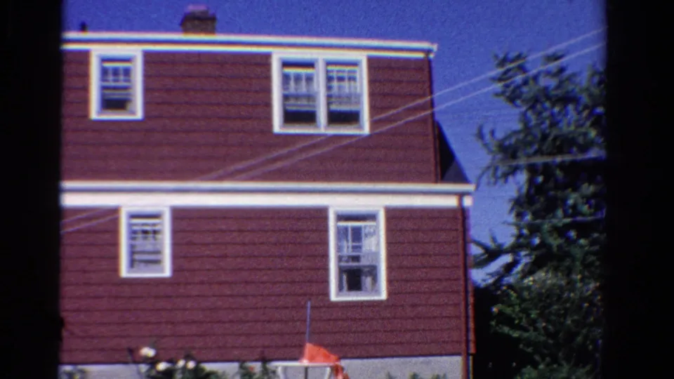 1958 Ontario home-movie frame showing the side of a red house with multiple white-trimmed windows and overhead lines.