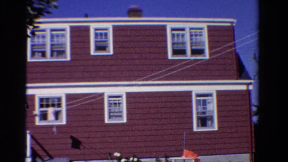 1958 Ontario home-movie frame of a red-sided house exterior with white-trimmed windows, overhead lines, and yard objects near the wall.