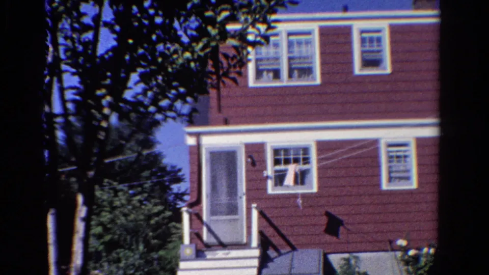 1958 Ontario 8mm still of a maroon two-story house with white trim, screened door, steps, clothesline, trees, and flowers.