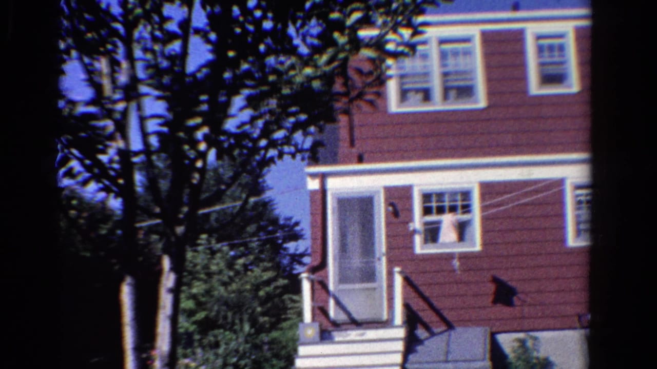 1958 Ontario home-movie view of a red-sided house exterior framed by trees, with screen doors, grass, and laundry on a line.