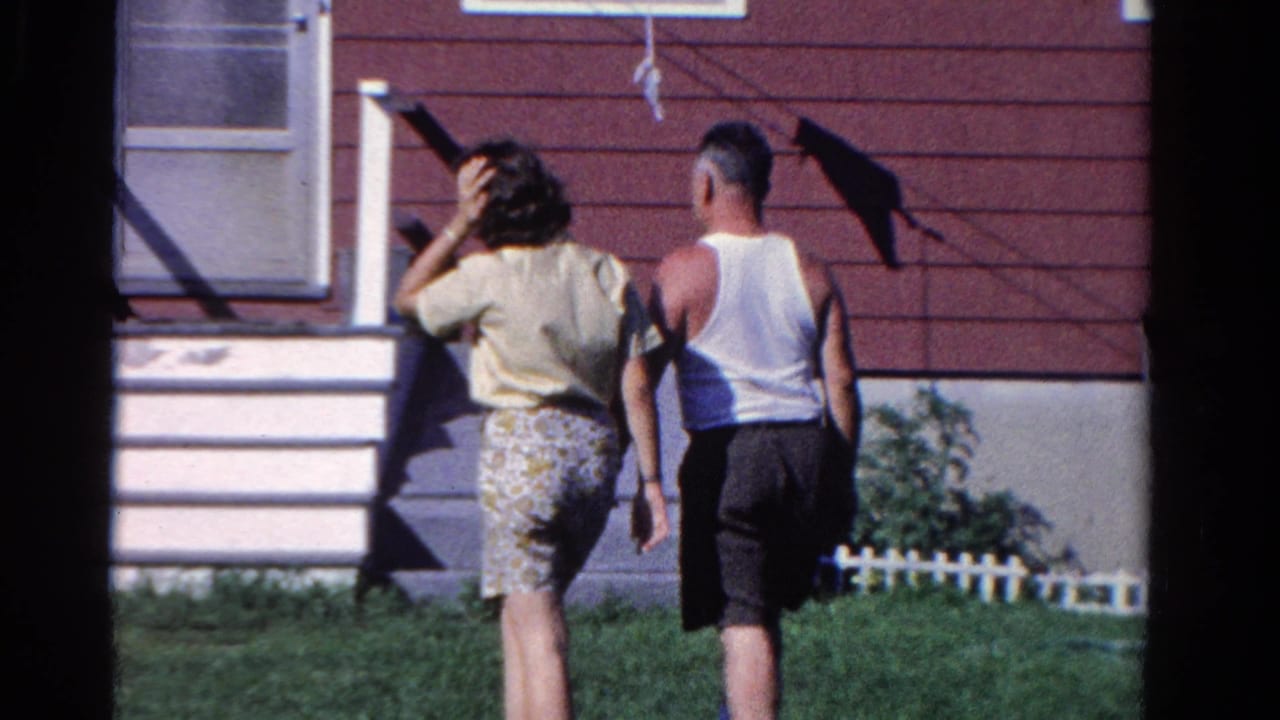 Two adults walk across a lawn toward the front steps of a red-sided house.