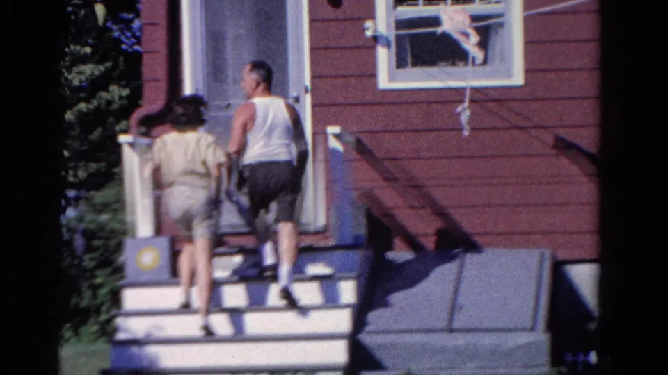 The woman and man continue up the porch steps, seen from behind beside the red house wall and window.