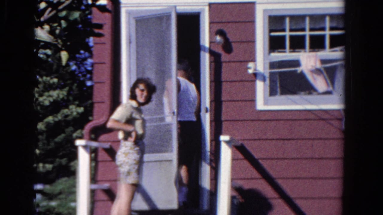 In 1958 Ontario home-movie footage, a woman stands on a porch outside a red-sided house while a man moves near the screen door.