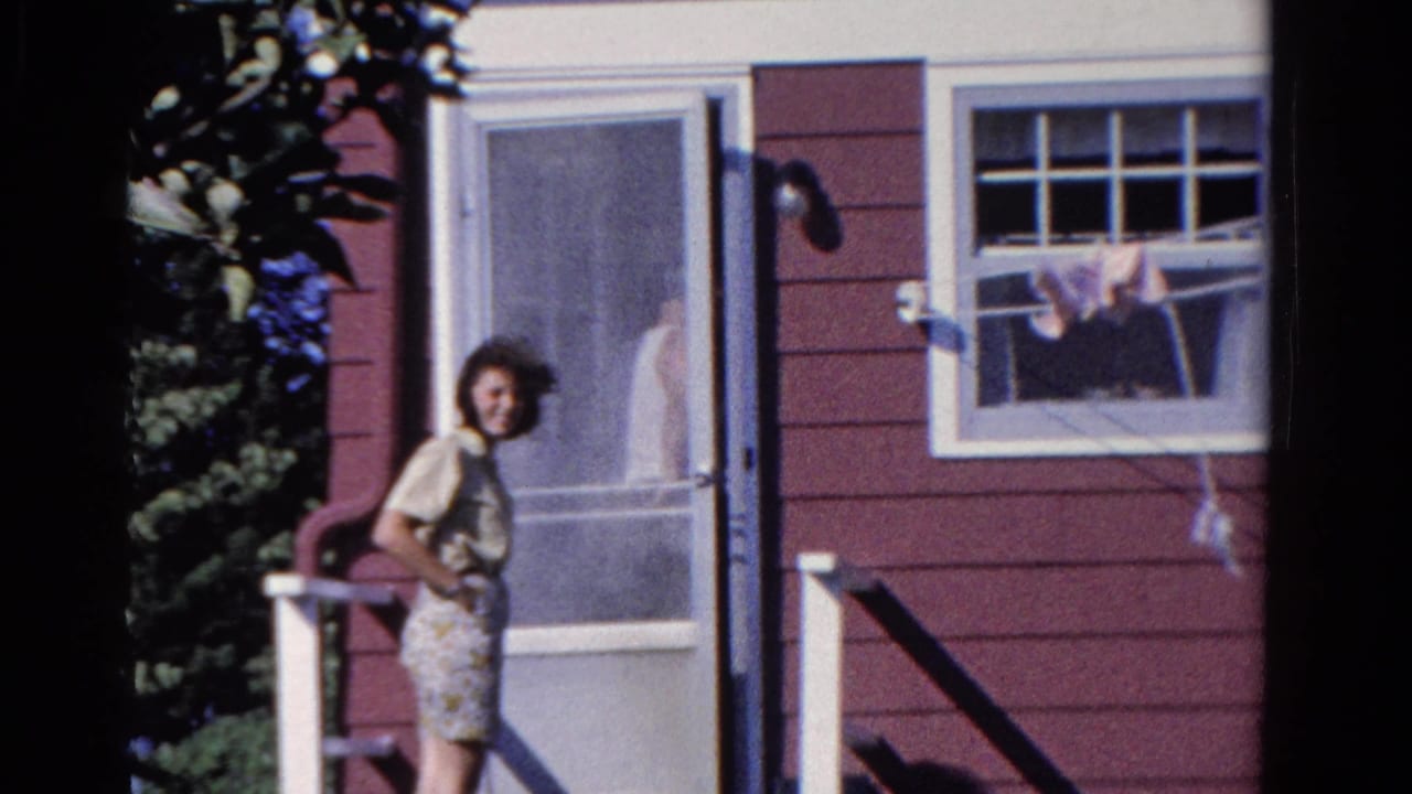Woman looks toward the camera beside a screen door, with a window and hanging laundry on the red house wall.