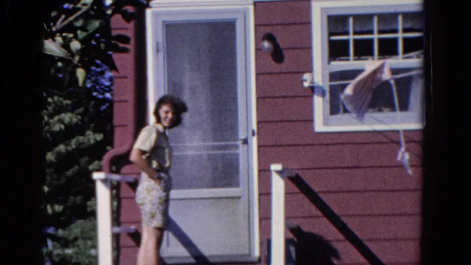 A woman remains on the porch outside the red-sided house in Ontario, framed by the screen door, porch railing, white trim, and nearby window.