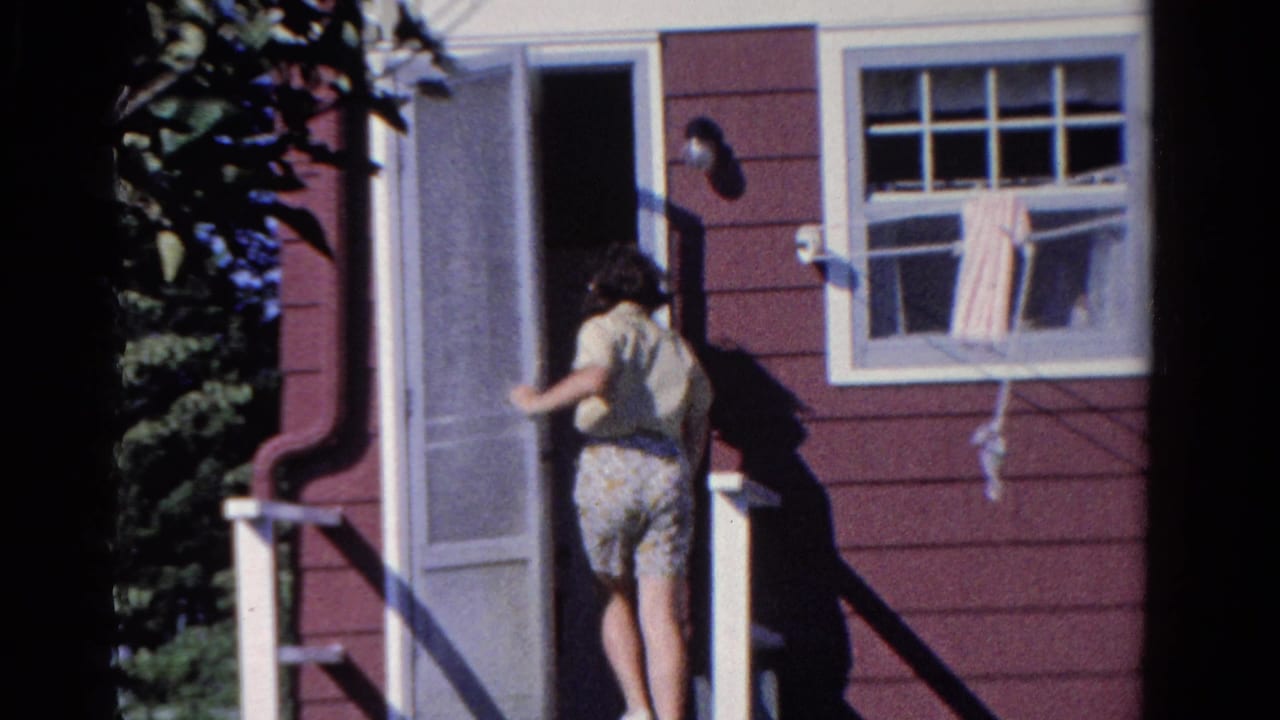 In a 1958 Ontario home-movie frame, a young woman approaches a white screen door on a red-sided house with a curtained window nearby.