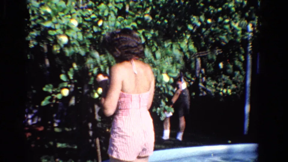 The young woman is seen from behind in a pink striped outfit as she stands close to the leafy fruit tree in the 1958 Ontario backyard scene.