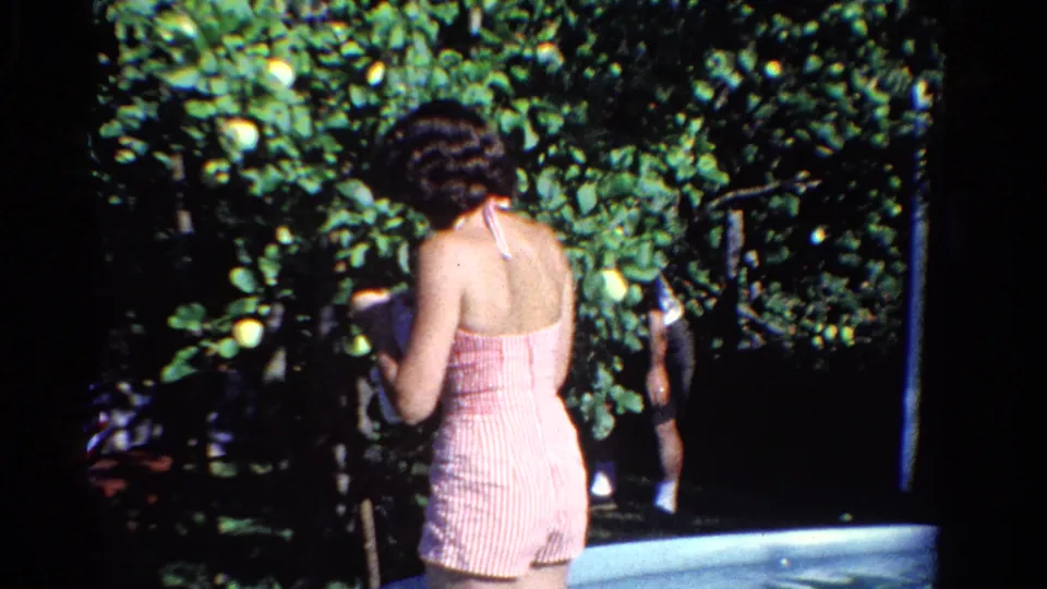 A final 1958 Ontario home-movie frame shows the young woman from behind near the fruit tree, with strong sunlight across the yard.