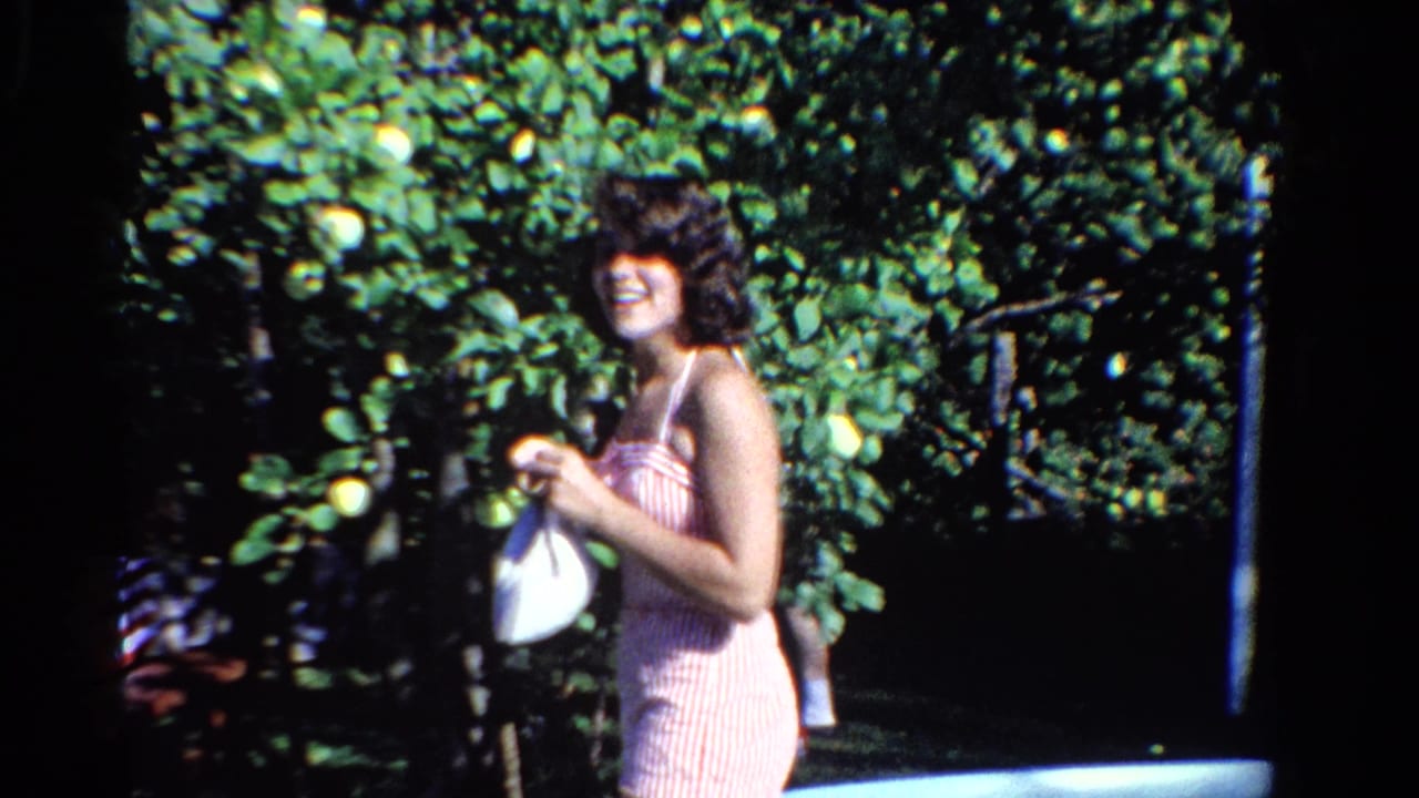 In Ontario, Canada, 1958, a woman in a pink striped swimsuit smiles beside a backyard pool while holding a white bag near leafy fruit trees.