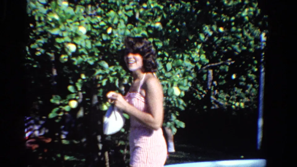 In Ontario, Canada, 1958, a woman in a pink striped swimsuit smiles beside a backyard pool while holding a white bag near leafy fruit trees.