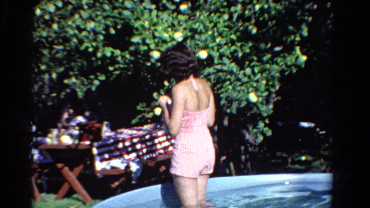Woman in a pink striped swimsuit stands in a backyard pool near a picnic table.