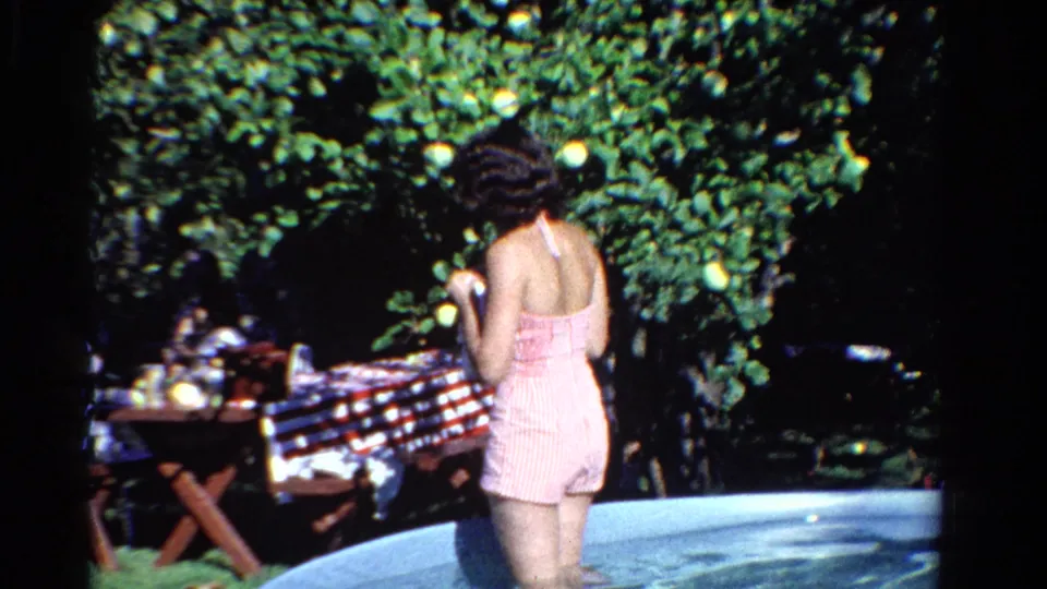 In Ontario, Canada, 1958, the woman is seen from behind in the pool near a picnic table covered with a red and white checkered cloth.
