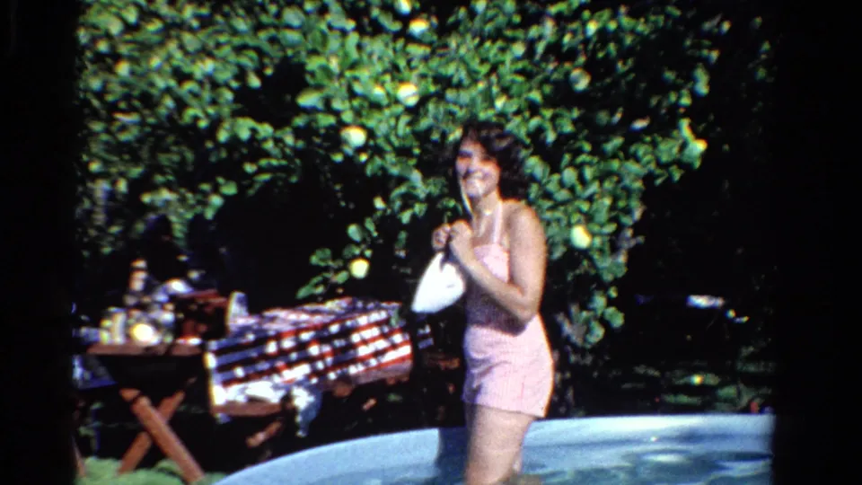 In Ontario, Canada, 1958, a woman in a pink striped swimsuit smiles from the backyard pool with a checkered picnic table behind her.